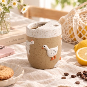 May include: A cylindrical ceramic mug with a speckled beige and white finish, featuring raised white goose figures with orange feet. The mug has a unique top design with a small circular opening and a larger crescent-shaped opening for drinking.