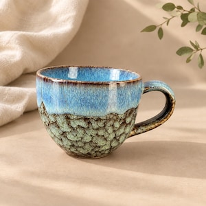 May include: A ceramic mug featuring a two-tone glaze with a smooth, bright blue upper section and a textured, moss-green lower section characterised by a crackled, crystalline pattern. The handle is dark brown with blue and green highlights.