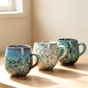 May include: Three ceramic mugs with rounded bodies and handles. The first mug features a blue and green drip glaze. The second mug has a white and grey crackle pattern. The third mug displays a dark green and black textured glaze finish.