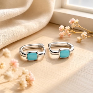 May include: A pair of silver hoop earrings featuring a square-shaped, smooth, bright turquoise stone inlay centred on the front of each hoop. The earrings have a polished metallic finish and a hinged closure mechanism for secure wear.