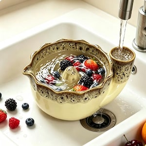 May include: A cream-coloured ceramic berry colander with a brown speckled rim and a built-in side funnel for water intake. The bowl is filled with water, fresh strawberries, blackberries, and blueberries, with a central drainage cone.