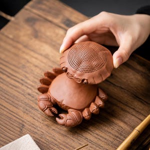 Puede incluir: Un colador de té de arcilla de color marrón rojizo con forma de cangrejo. El caparazón superior es extraíble y presenta una malla fina de pequeñas perforaciones circulares para filtrar las hojas de té. La base está esculpida con patas y pinzas.