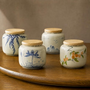 May include: Four small ceramic jars with light-colored wooden lids. Each jar features a unique hand-painted design: one with blue willow branches, one with a blue lotus flower, one with blue mountain silhouettes, and one with orange fruit branches.