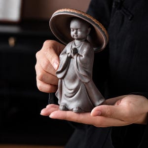 Peut inclure: Une petite figurine en céramique gris foncé représentant un moine aux yeux fermés, les mains jointes en prière. Le personnage porte une robe et est abrité par un grand chapeau texturé en forme de champignon brun qui recouvre sa tête.