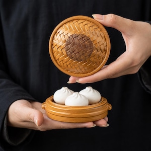 Op de afbeelding: Een kleine, ronde, lichtbruine bamboe stoommand met een gevlochten deksel met een donker cirkelvormig ontwerp in het midden. In de basis zijn drie gladde, witte, ronde, knoedelvormige objecten in een driehoekige formatie gerangschikt.
