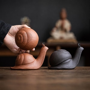 May include: Two snail-shaped ceramic tea strainers, one in reddish-brown and one in dark charcoal grey. Each features a textured shell with a perforated top section for straining, a smooth body, and two small antennae protruding from the head.