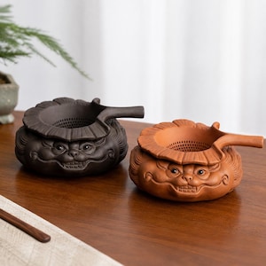 Peut inclure: Deux passoires à thé rondes en céramique avec poignées, l'une brun foncé et l'autre orange terre cuite. Chaque passoire présente un visage sculpté aux dents apparentes et un large bord froncé rappelant une feuille. L'intérieur est perforé.