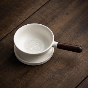 Op de afbeelding: Een witte keramische theezeef met een ronde kom en een geperforeerde bodem voor het filteren. De zeef is voorzien van een lange, cilindrische donkerbruine houten handgreep aan de zijkant, wat een contrast vormt met de matte witte afwerking.