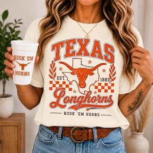May include: A cream-colored t-shirt featuring a graphic with the text TEXAS, Longhorns, and HOOK 'EM HORNS, alongside a longhorn skull, a map of Texas, and a checkered pattern in orange. A white cup with the text HOOK 'EM HORNS and a longhorn icon.