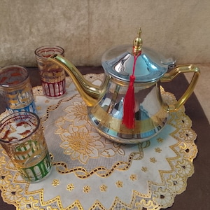 May include: A polished silver and gold-toned metal teapot with a long curved spout, a handle, and a lid topped with a small finial. A bright red tassel hangs from the lid. Two clear glass tumblers feature gold and colored decorative patterns.