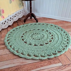 May include: A round, sage green crocheted rug featuring a detailed, openwork circular pattern with a central starburst design and a scalloped edge. The textile is crafted from yarn with a visible, textured stitch throughout the entire piece.