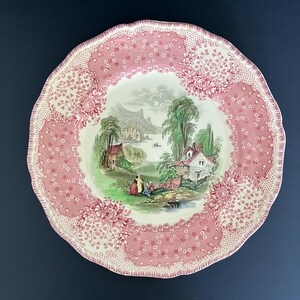 May include: A round ceramic plate with a scalloped edge featuring a central landscape illustration of a lake, trees, buildings, and figures. The border is decorated with a pink and white floral pattern with intricate stippled details.