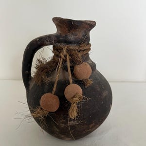 May include: A dark brown, round clay pitcher with a single handle and a wide neck. The neck is wrapped with natural fiber twine, which features three dangling, spherical, light brown clay beads attached to the ends of the twine strands.