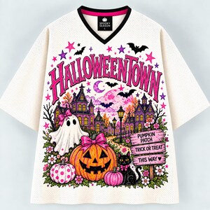 May include: A cream-colored V-neck jersey featuring a graphic print of Halloweentown with a ghost, a jack-o'-lantern, a black cat, and a signpost reading Pumpkin Patch, Trick or Treat, and This Way, accented with pink stars and bats.