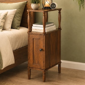 May include: A tall, narrow wooden nightstand with a dark brown finish, featuring a single cabinet door with a round black knob, four turned legs, and an open top shelf. The shelf holds a black metal lantern, a digital clock, and three stacked books.