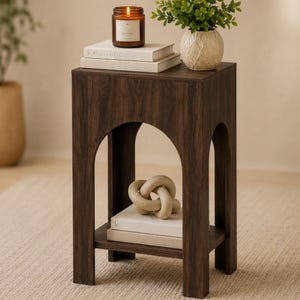 May include: A dark brown wood-grain side table featuring an arched cutout design on the front and sides. The table has a flat top surface and a lower shelf. The top holds two stacked white books and a lit candle, while the shelf holds a beige knot decor.