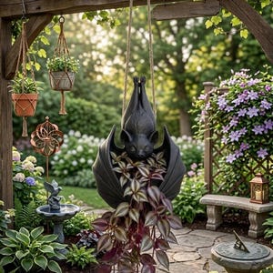 May include: A black bat-shaped hanging planter suspended by a thick rope. The bat is depicted in a resting position with wings wrapped around a trailing plant featuring variegated purple and green leaves. The planter is made of a dark material.