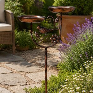 Könnte beinhalten: Eine kupferfarbene Vogeltränke aus Metall mit zwei runden Becken auf unterschiedlichen Höhen, die von einem zentralen Stab getragen werden. Die Struktur ist mit einem geformten Metallvogel, Rankenmotiven und einer Blume am Stiel verziert.