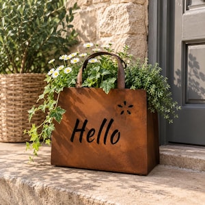 May include: A rectangular, rust-coloured metal planter with a handle, featuring the word Hello cut out in black lettering and a small flower-shaped cutout. The planter is filled with green foliage and white daisy-like flowers.