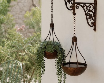 May include: A dark brown, rusted metal wall-mounted bracket with two ornate, scrolling arms. Two matching metal bowls hang from the bracket by chains. One bowl contains a green succulent plant with trailing foliage, while the other holds water.