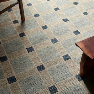 May include: A woven area rug featuring a grid pattern of large light blue and beige squares, accented by small dark navy blue squares at the intersections. The rug has a textured, low-pile surface and covers the floor area between two wooden furniture legs.