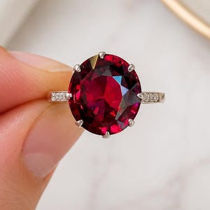 Op de afbeelding: Een ring met een grote, ovaal geslepen, dieprode edelsteen in een zilverkleurige metalen band. De band is aan de zijkanten versierd met kleine, heldere, fonkelende steentjes, vastgezet in een zetting die de centrale rode steen vasthoudt.