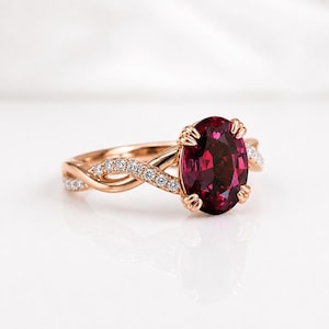 Könnte beinhalten: Ein Ring aus Roségold mit einem großen, oval geschliffenen, tiefroten Edelstein, der von vier Krappen gehalten wird. Die Ringschiene hat ein gedrehtes Design und ist mit kleinen, runden, klaren Steinen besetzt, die entlang der Schiene verlaufen.