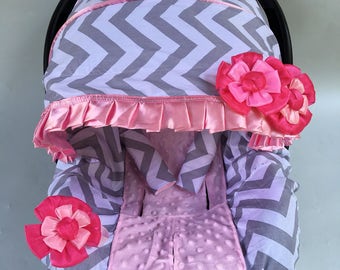May include: A gray and pink chevron car seat cover with a pink and white ruffled trim and pink flower accents.