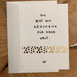 Könnte beinhalten: Eine weiße Grußkarte mit passendem Umschlag, die den handgeschriebenen schwarzen Text DU BIST EIN GESCHENK FÜR DIESE WELT zeigt. Unter dem Text befindet sich ein horizontaler Streifen mit gold-braunem Punktemuster und ein kleines schwarzes Herz.