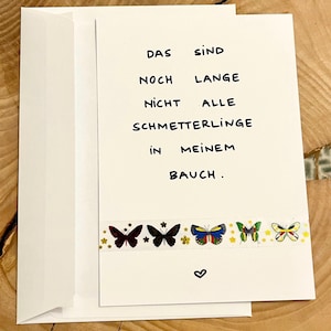 Könnte beinhalten: Eine weiße Grußkarte mit passendem Umschlag, bedruckt mit dem Text DAS SIND NOCH LANGE NICHT ALLE SCHMETTERLINGE IN MEINEM BAUCH. Unter dem Text befindet sich ein horizontaler Streifen aus Dekoband mit bunten Schmetterlingsillustrationen.