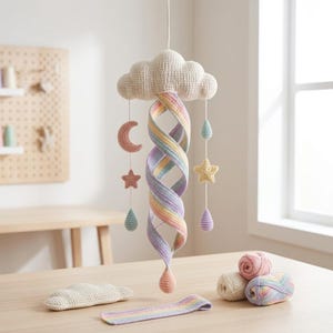 May include: A crochet mobile featuring a central cream-colored cloud with a hanging spiral rainbow ribbon. Suspended from the cloud are small crocheted shapes including a pink crescent moon, a yellow star, and pastel-colored raindrops.