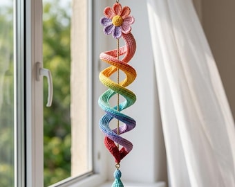 May include: A hanging crochet spiral ornament featuring a rainbow color gradient. The top is adorned with a multi-colored flower, and the bottom features a small, light blue teardrop-shaped charm. The entire piece is suspended by a thin gold chain.