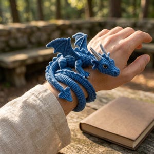 Puede incluir: Una figura de dragón tejida a ganchillo en color azul con una cola larga y enroscada que se envuelve alrededor de la muñeca. El dragón presenta alas texturizadas, dos pequeños cuernos grises en la cabeza y ojos oscuros de hilo azul.