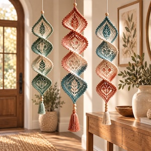 May include: Three vertical hanging decorations featuring spiraling crochet motifs in shades of sage green, cream, terracotta, and blue. Each piece consists of four connected diamond-shaped panels with leaf patterns, finished with wooden beads and tassels.
