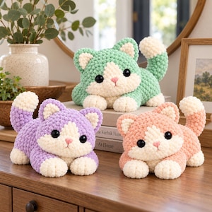 May include: Three plush crochet cats in mint green, lavender, and peach. Each cat features a cream-colored face, chest, and paws, with large black plastic eyes and a small pink stitched nose. The yarn has a soft, textured, bobble-like appearance.