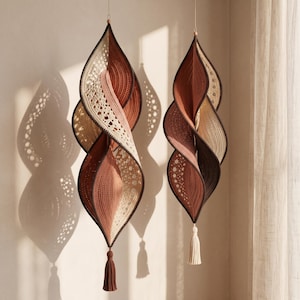 May include: Two hanging spiral ornaments crafted from crocheted fabric in shades of cream, terracotta, and dark brown. Each piece features an elongated, twisted leaf-like shape with intricate openwork patterns and a single tassel at the bottom.