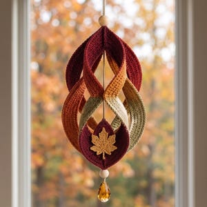 May include: A hanging crochet ornament featuring twisted loops in shades of burgundy, orange, and olive green. A small crocheted maple leaf is attached to the center, accented by two light wooden beads and a faceted amber-colored crystal drop.