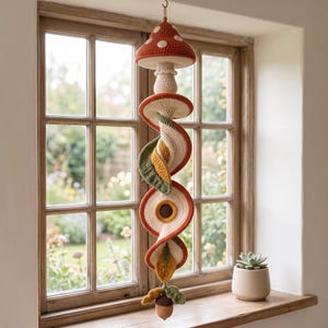 May include: A hanging crochet mobile featuring a top mushroom cap with white spots, followed by a series of spiralling fabric elements in shades of orange, cream, olive green, and mustard yellow, ending with a small brown acorn and green leaves.