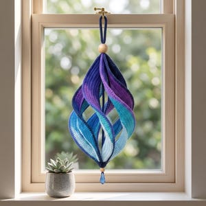 May include: A hanging crochet ornament featuring a spiraling, three-dimensional shape with a color gradient transitioning from deep navy blue at the top to vibrant purple, teal, and light blue at the bottom, accented by two wooden beads and a blue crystal.