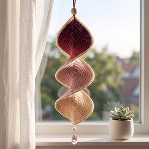 May include: A hanging crochet spiral ornament featuring a gradient color scheme transitioning from deep burgundy at the top to light pink in the middle and cream at the bottom. It is suspended by a string with a wooden bead and a clear crystal drop.