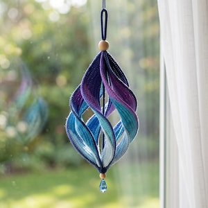 May include: A hanging ornament featuring a spiraling, multi-colored crochet design in shades of dark blue, purple, teal, and light blue. The structure is suspended by a dark blue cord with a wooden bead at the top and a blue crystal drop at the base.