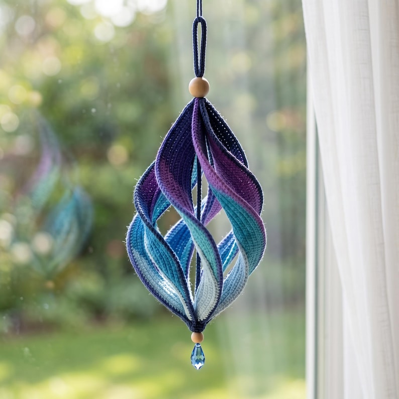 May include: A hanging ornament featuring a spiraling, multi-colored crochet design in shades of dark blue, purple, teal, and light blue. The structure is suspended by a dark blue cord with a wooden bead at the top and a blue crystal drop at the base.