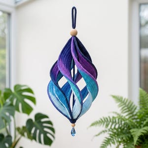 Può includere: Un ornamento a spirale in uncinetto da appendere con una sfumatura di colori che passa dal blu navy profondo al viola, turchese e azzurro. La struttura è sormontata da una perla di legno e termina con un pendente in cristallo blu.