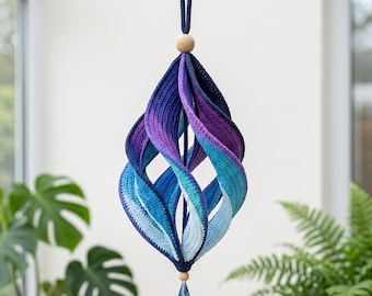 Könnte beinhalten: Ein hängendes Häkel-Spiralornament mit einem Farbverlauf von tiefem Marineblau zu leuchtendem Violett, Türkis und Hellblau. Die Struktur ist mit einer Holzperle versehen und endet in einem blauen Kristallanhänger an der Unterseite.