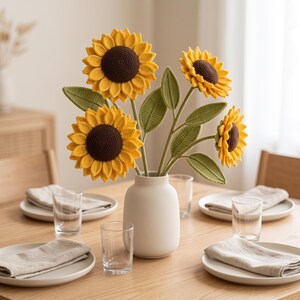 Könnte beinhalten: Ein Strauß aus vier gehäkelten Sonnenblumen mit leuchtend gelben Blütenblättern und dunkelbraunen Mitten, ergänzt durch strukturierte grüne Blätter an den Stielen. Die Blumen stehen in einer glatten, matten, cremeweißen Keramikvase.