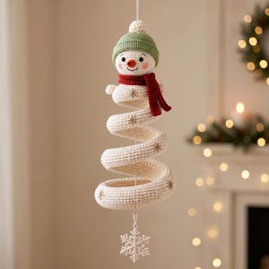 May include: A crocheted hanging ornament featuring a snowman head with a green beanie and red scarf, attached to a long, cream-colored spiral body decorated with small embroidered gold snowflakes, ending with a white plastic snowflake charm.