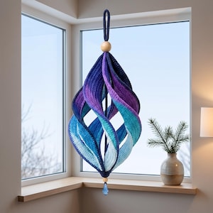 May include: A hanging spiral ornament crafted from crocheted yarn in a gradient of colors transitioning from deep navy blue to purple, teal, and light blue. It features a wooden bead at the top and bottom, with a blue crystal drop at the base.