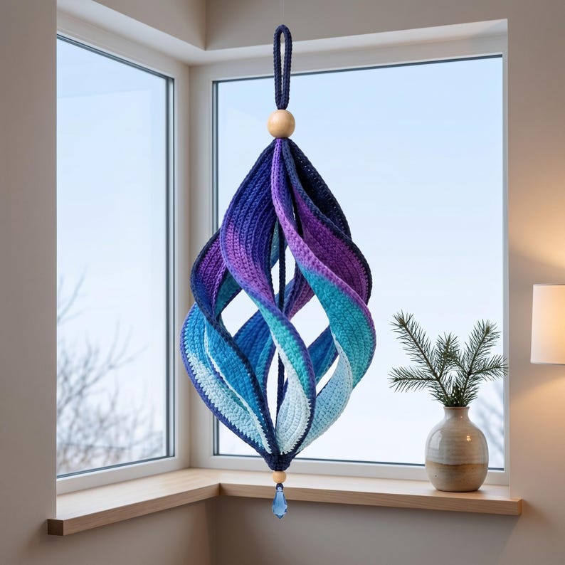May include: A hanging spiral ornament crafted from crocheted yarn in a gradient of colors transitioning from deep navy blue to purple, teal, and light blue. It features a wooden bead at the top and bottom, with a blue crystal drop at the base.