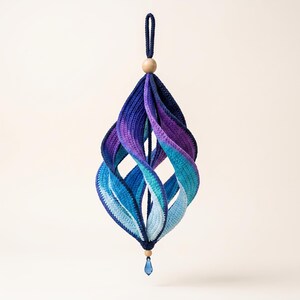 May include: A hanging crochet ornament featuring a spiraling, multi-layered design in a gradient of colors transitioning from deep navy blue to purple, teal, and light blue. It is topped with a wooden bead and finished with a small blue crystal.