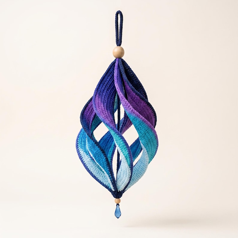 May include: A hanging crochet ornament featuring a spiraling, multi-layered design in a gradient of colors transitioning from deep navy blue to purple, teal, and light blue. It is topped with a wooden bead and finished with a small blue crystal.